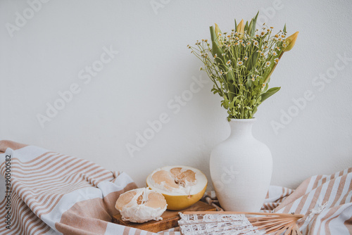 rustical still life with pomelo and fresh simple bouquet of yellow tulips and wild camomile and laced hand fan, white background, space for text photo