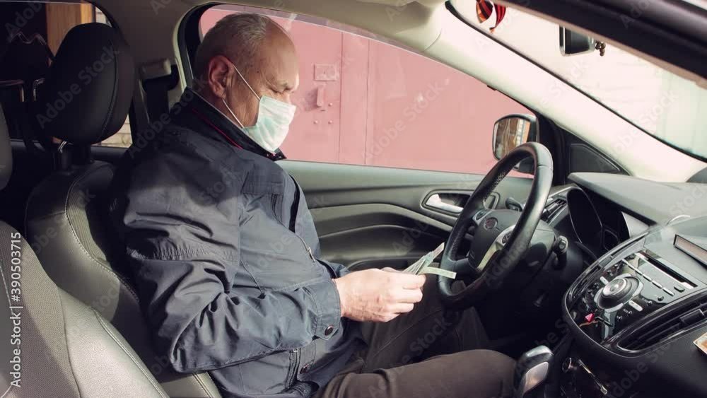 A man in a mask on his face sitting in a car is counting money. The vehicle is at a standstill. Against the background of a brown wall. Warm, autumn day. Soft light. Close-up. UHD. A man in a mask on his face sitting in a car is counting money. The vehicle is at a standstill. Against the background of a brown wall. Warm, autumn day. Soft light. Close-up. UHD.