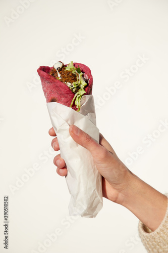 mano femenina sosteniendo falafel en pan de pita de remolacha con salsa de lima, cilantro y yogur en papel de mantequilla blanca envoltura de fondo blanco