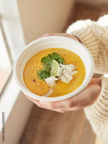 Vista aérea de manos femeninas sosteniendo sopa de batata con leche de coco, semillas, perejil, coco, fondo de madera de colores cálidos a lado de una ventana luz natural