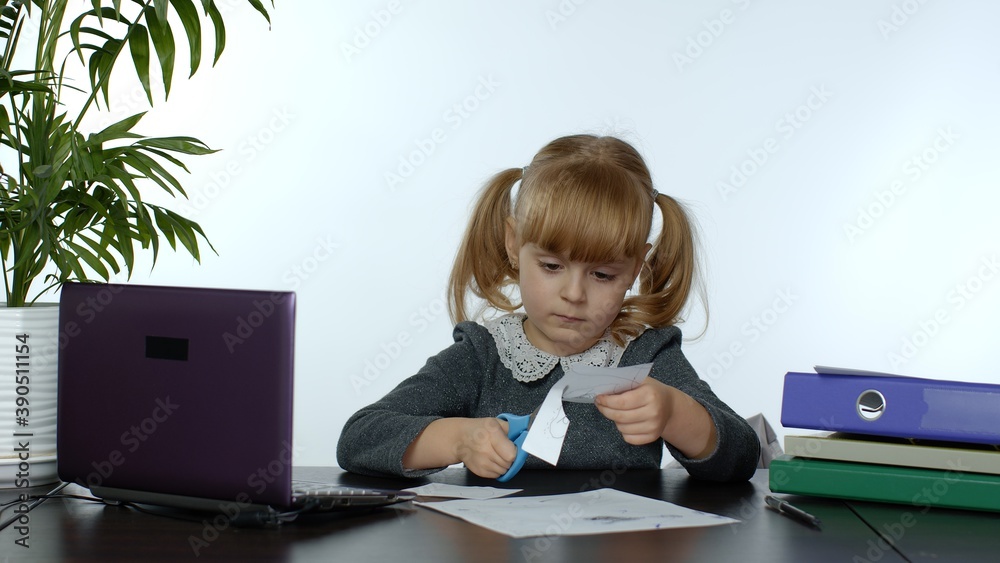 Child schoolgirl learns lessons at home sitting at table cutting with scissors shapes out of paper. Kid girl distance education. Studying during coronavirus quarantine. White background Child schoolgirl learns lessons at home sitting at table cutting with scissors shapes out of paper. Kid girl distance education. Studying during coronavirus quarantine. White background