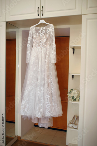 A white wedding dress is hung on a hanger next to the wedding accessories, shoes and the bride's bouquet. The bride is ready to put on her outfit. Wedding accessories concept for a perfect wedding
