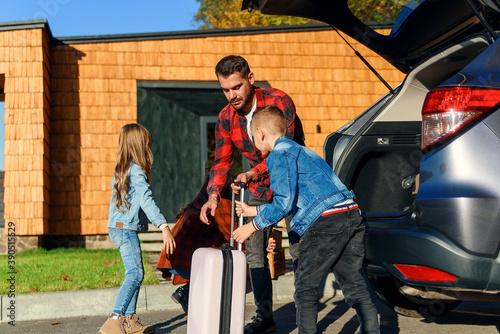 A happy family unpacks luggage from the trunk of a car after moving to a new home.