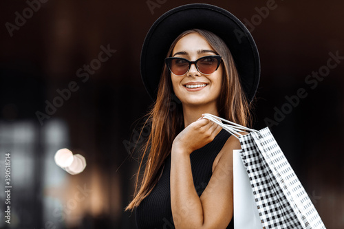 attractive young woman in a black dress and hat, dark glasses, holding shopping bags, enjoying a successful shopping, in the background of the shopping center. Black Friday, shopping