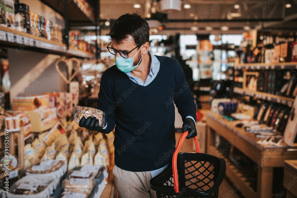 Casually dressed middle age man with face protective mask and gloves buying healthy food and drink in a modern supermarket or grocery store. Pandemic or epidemic lifestyle and consumerism concept.
