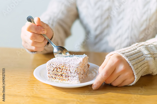 Women's hands hold a teaspoon and cut a piece of delicious cake in a cafe on a brown wooden table. Cup of coffee on the table. Close-up, selective focus on the spoon