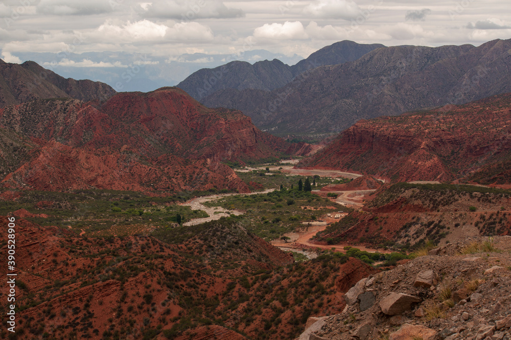 Geology. Panorama view of the red sandstone and rocky mountains, green forest and river flowing across the valley under a cloudy sky in Miranda Slope, La Rioja Argentina.