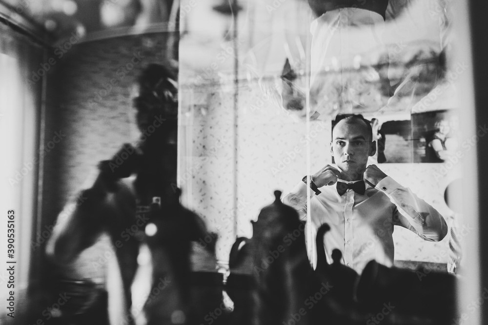 The man looks at his reflection in the mirror and straightens his bow tie on shirt at home. clothing concept. Black and white photo.