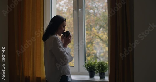 Side portrait of pensive girl standing by window with cup of tea slow motion. Young woman drinking hot coffee looking outside at autumn landscape copy space. Loneliness depression solitude concept