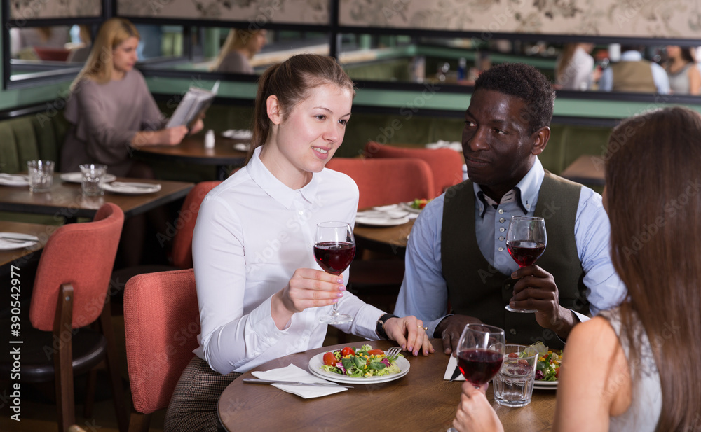 Young happy couple on friendly meeting with female friend over dinner with wine in restaurant