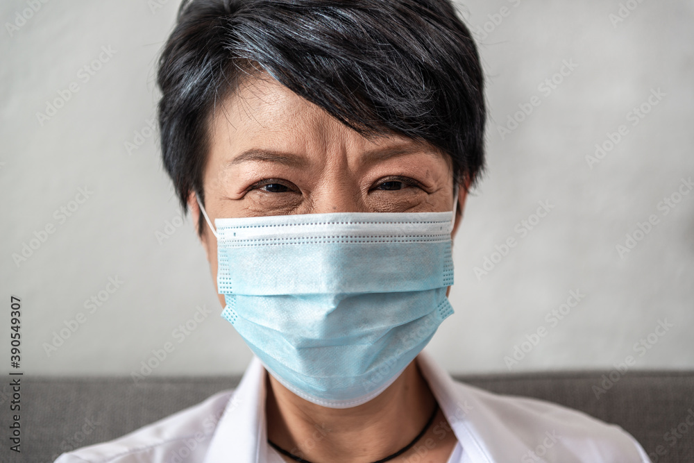 A close-up of happy Aged Asian woman with gray hair smile under a sanitary mask on her face against coronavirus or COVID-19 disease sitting on a couch at home. And the positive Elderly concept
