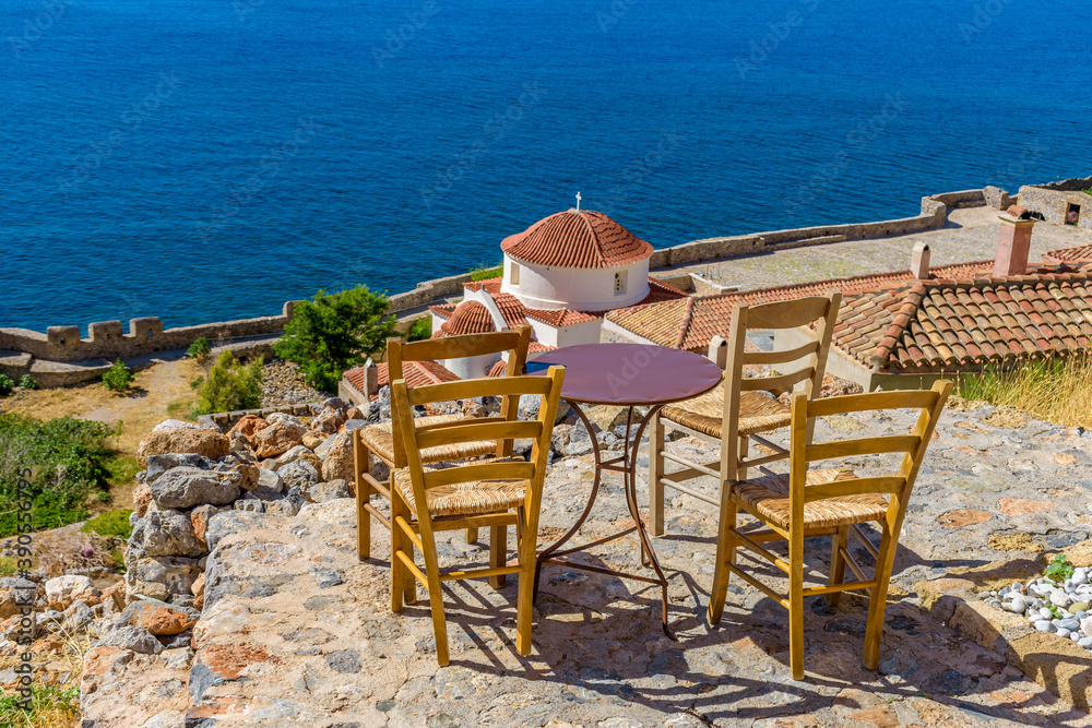Traditional cafe exterior in the fortified medieval  castle of Monemvasia. Iron tables and wooden chairs with the view of the church panagia chrysafitissa and the aegean sea in the background.
