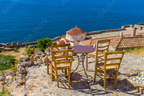 Traditional cafe exterior in the fortified medieval  castle of Monemvasia. Iron tables and wooden chairs with the view of the church panagia chrysafitissa and the aegean sea in the background.