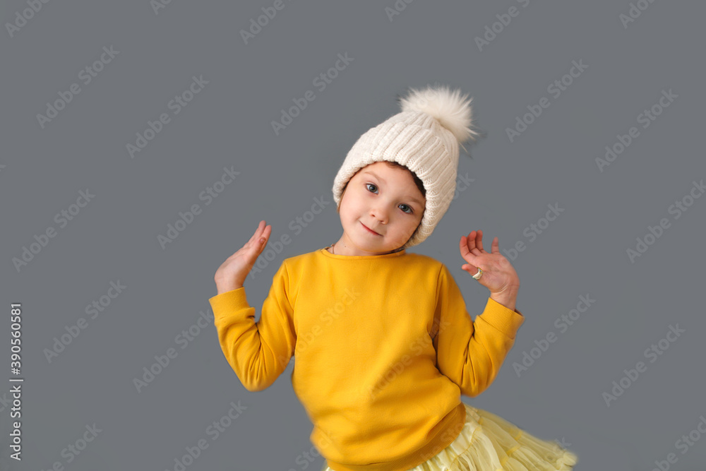 Funny crazy caucasian child girl in white knitted hat and yellow sweater on a gray background, the concept of childish emotions and antics. Funny crazy caucasian child girl in white knitted hat and yellow sweater on a gray background, the concept of childish emotions and antics.