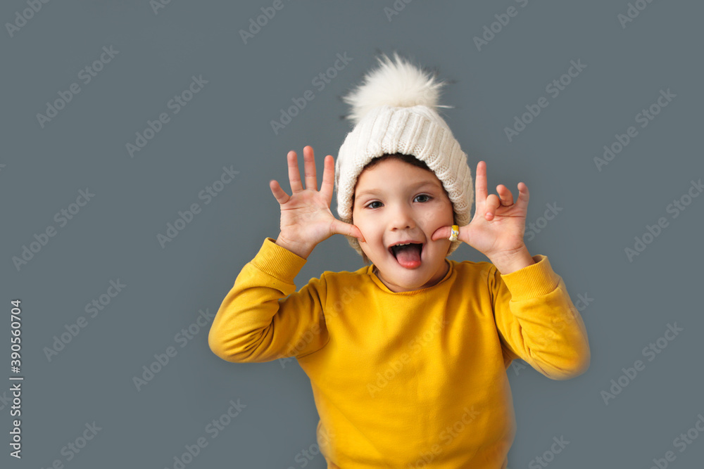 Funny crazy caucasian child girl in white knitted hat and yellow sweater on a gray background, the concept of childish emotions and antics. Funny crazy caucasian child girl in white knitted hat and yellow sweater on a gray background, the concept of childish emotions and antics.