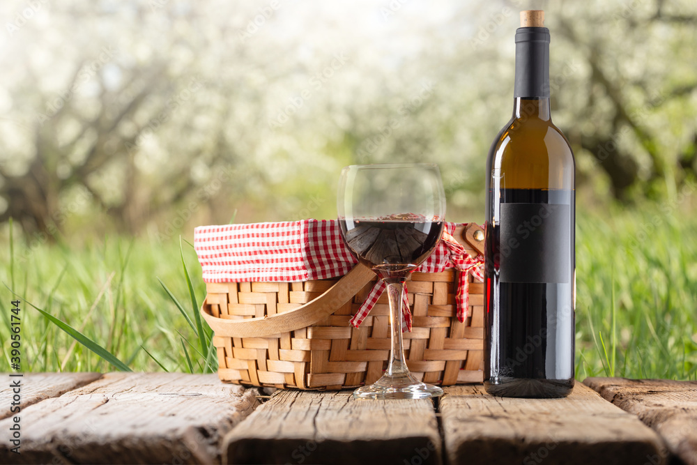 Picnic basket with red wine bottle and wine glass stands on wooden rustic table on green garden background. Concept of romantic weekends. Tasty alcoholic drink. Picnic basket with red wine bottle and wine glass stands on wooden rustic table on green garden background. Concept of romantic weekends. Tasty alcoholic drink.