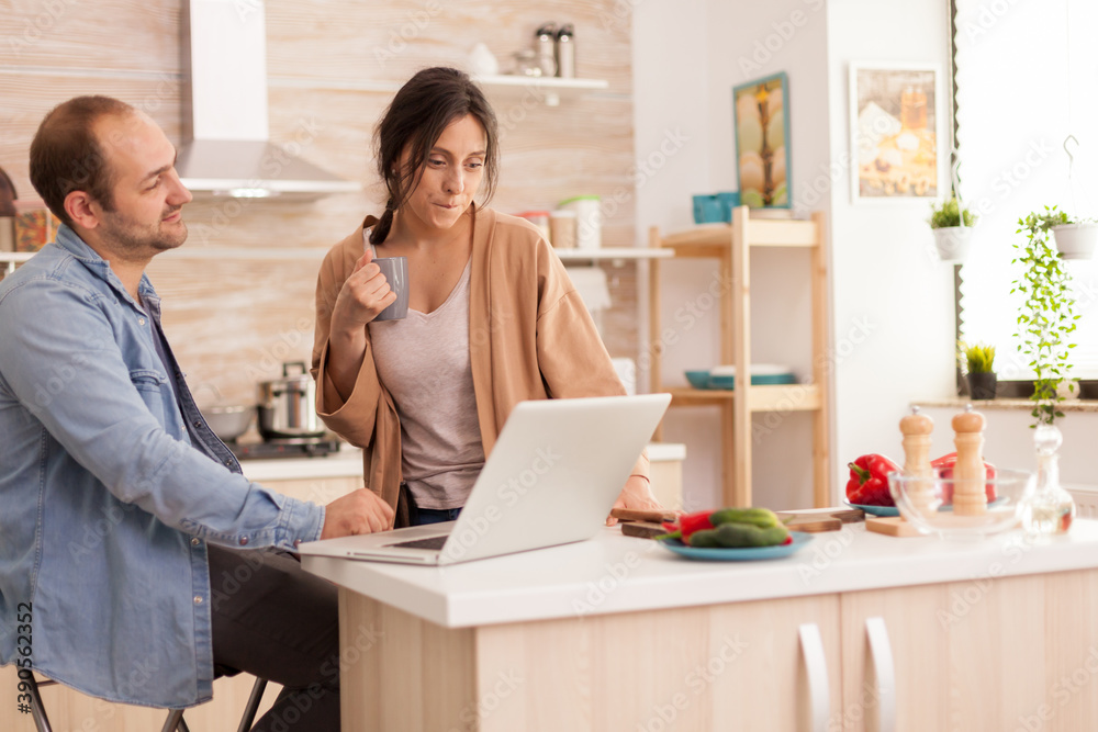 Woman looking at husband laptop while he is working in kitchen. Girlfriend holding cup of coffee. Happy loving cheerful romantic in love couple at home using modern wifi wireless internet technology