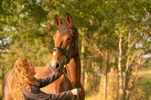 A brown horse head, with a bridle, woman next to the horse, in the autumn evening sun