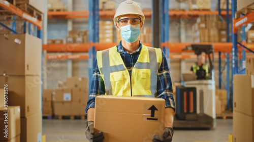 Handsome Male Worker Wearing Medical Face Mask and Hard Hat Carries Cardboard Box Stands in Retail Warehouse full of Shelves with Goods. Logistics, Distribution Center. Fromt Shot photo
