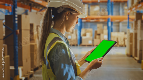 Professional Female Worker Wearing Hard Hat Uses Digital Tablet Computer with Green Chroma Key Screen in Portrait Mode in the Retail Warehouse full of Shelves with Goods. Logistics and Distribution 