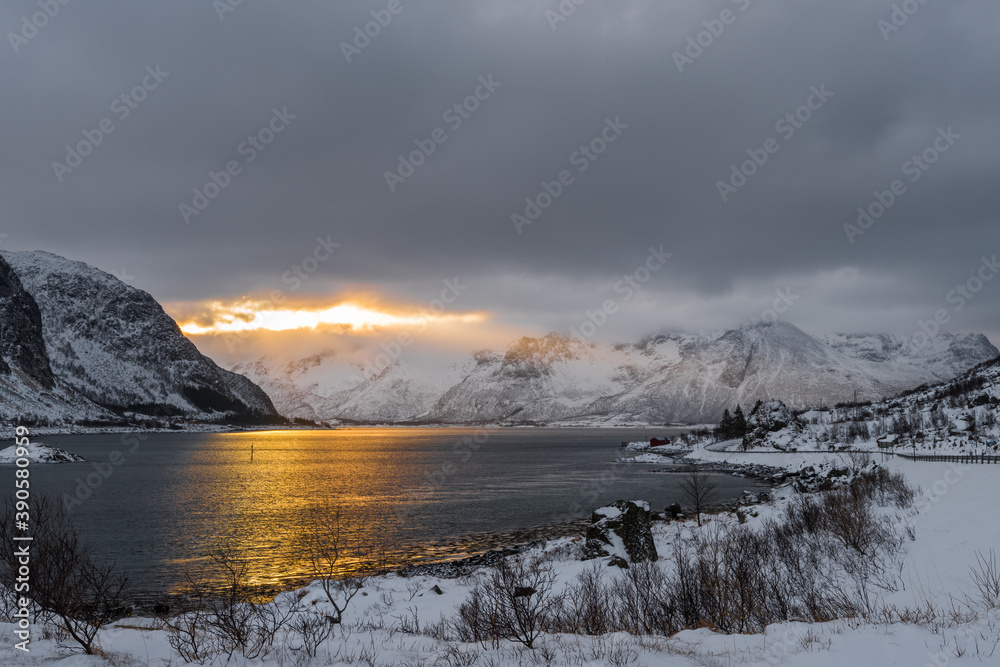 Scenic view over morning sun breaking through dense clouds behind snow-clad mountains near lake on the Lofoten islands archipelago in Norway