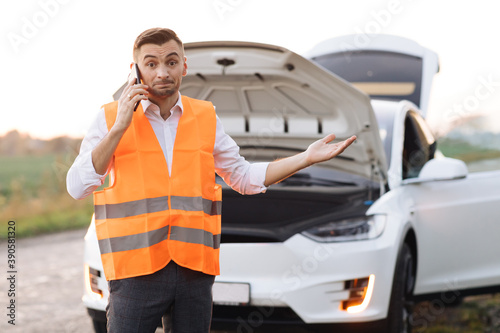 Man calling car assistance services because his electric car is broken. Concept road accident. Help repair. Man in a safety vest talking on cell phone