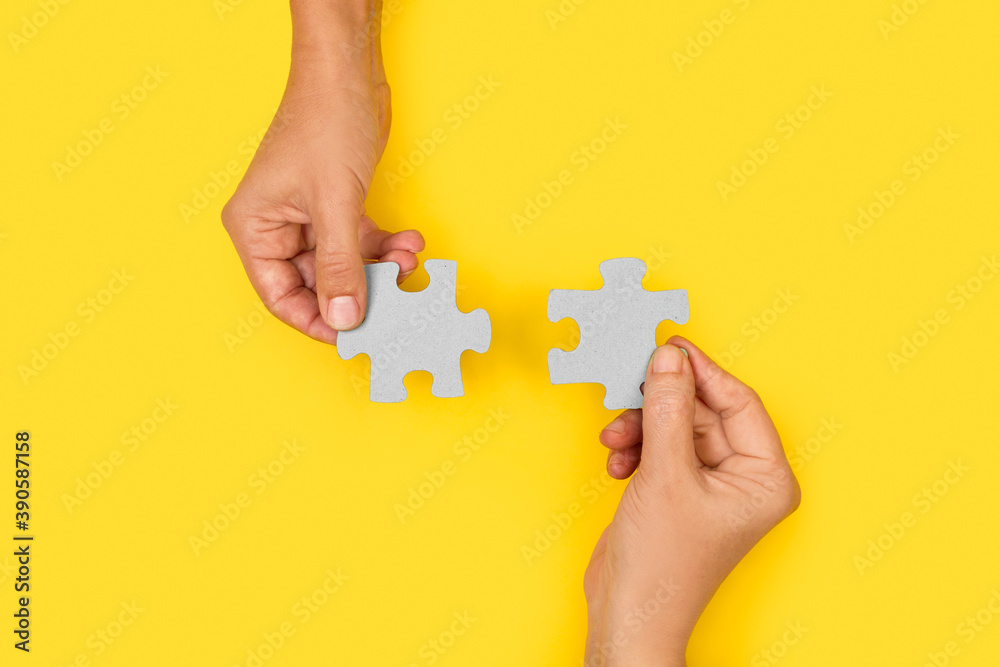 Manos uniendo dos piezas de puzzle sobre un fondo amarillo liso y aislado. Vista superior. Copy space - obrazy, fototapety, plakaty 