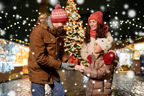 family, winter holidays and celebration concept - happy mother, father and little daughter with gift at christmas market on town hall square in tallinn, estonia over snow