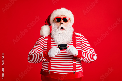Portrait of his he nice handsome amazed stunned wondered bearded fat Santa holding in hand bank card modern novelty service reaction isolated over bright vivid shine vibrant red color background © deagreez