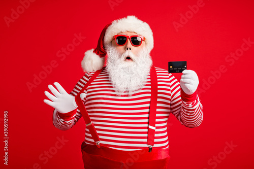 Portrait of his he nice handsome amazed stunned Santa using holding in hand bank card December bargain pulling suspender omg wow isolated over bright vivid shine vibrant red color background © deagreez