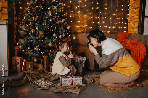 Mother taking pictures of her daughter at home, next to a christmas tree.