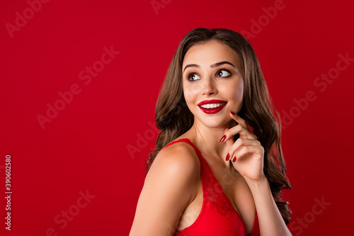 Photo portrait of hopeful woman looking behind shoulder at blank space touching cheek with fingers isolated on vivid red colored background