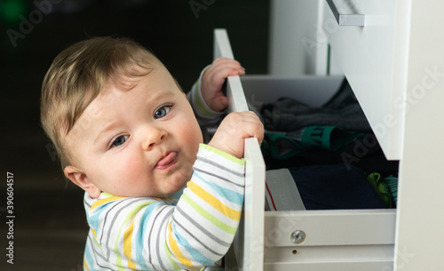 Obraz na płótnie Little caucasian baby boy opening a white drawer with underwear when nobody is watching. Naughty toddler exploring dangerous furniture. Baby safe at home concept. w salonie