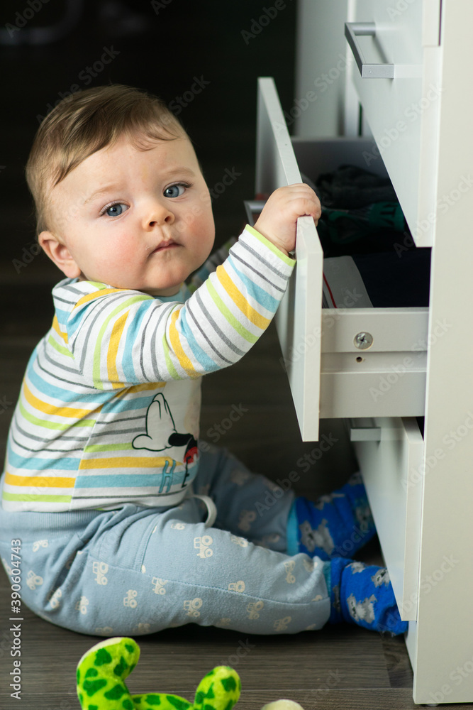 Little caucasian baby boy opening a white drawer with underwear when nobody is watching. Naughty toddler exploring dangerous furniture. Baby safe at home concept.