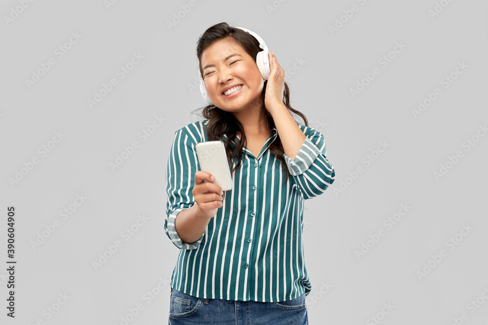people, technology and audio equipment concept - happy smiling asian young woman in headphones listening to music on smartphone over grey background