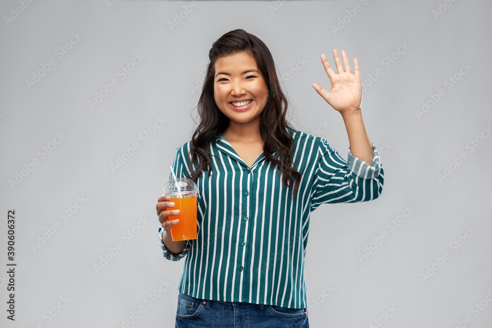 takeaway drinks and people concept - happy smiling young asian woman drinking orange juice from plastic cup with paper straw waving hand over grey background takeaway drinks and people concept - happy smiling young asian woman drinking orange juice from plastic cup with paper straw waving hand over grey background