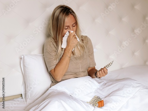 A woman with a cold sits on the bed, wipes her nose with a handkerchief and looks at the pills.