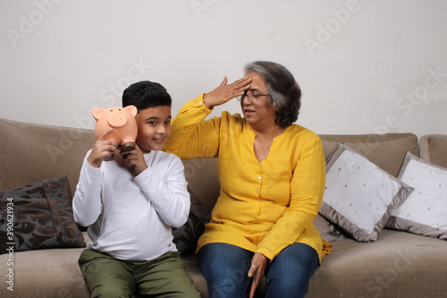 Fotografía Indian/asian Grandma teaching her grand son importance of savings, putting coins into pink piggy bank