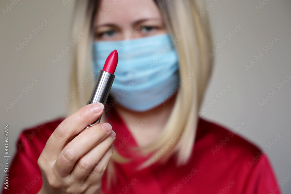 Woman in a protective medical mask with red lipstick in her hand. Makeup and cosmetics during the coronavirus pandemic Woman in a protective medical mask with red lipstick in her hand. Makeup and cosmetics during the coronavirus pandemic