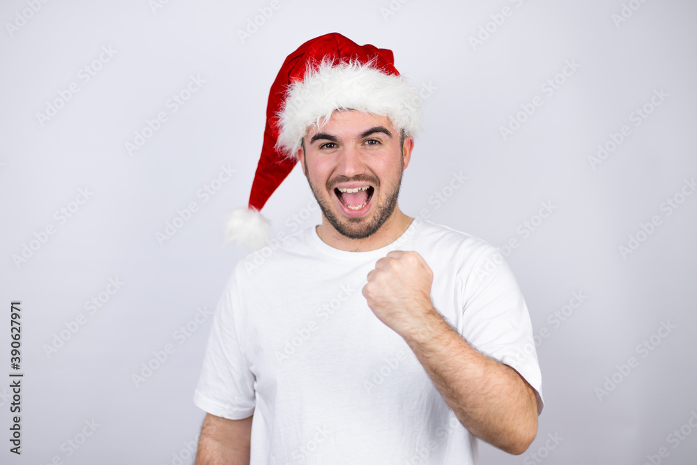Young handsome man wearing a Santa hat over white background angry and mad raising fist frustrated and furious while shouting with anger. Rage and aggressive concept. Young handsome man wearing a Santa hat over white background angry and mad raising fist frustrated and furious while shouting with anger. Rage and aggressive concept.