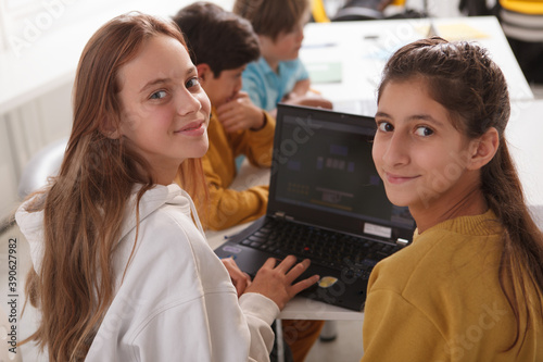 Happy teenage girls smiling to the camera while working on a school project together, using laptop