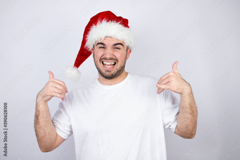Young handsome man wearing a Santa hat over white background shouting with crazy expression doing rock symbol with hands up Young handsome man wearing a Santa hat over white background shouting with crazy expression doing rock symbol with hands up