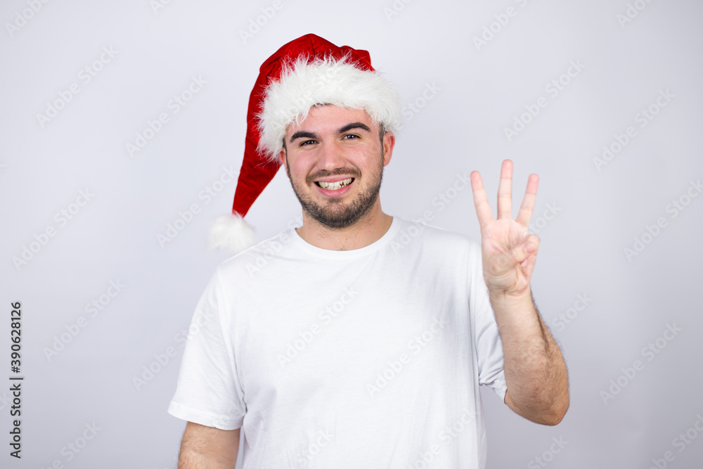 Young handsome man wearing a Santa hat over white background showing and pointing up with fingers number three while smiling confident and happy Young handsome man wearing a Santa hat over white background showing and pointing up with fingers number three while smiling confident and happy