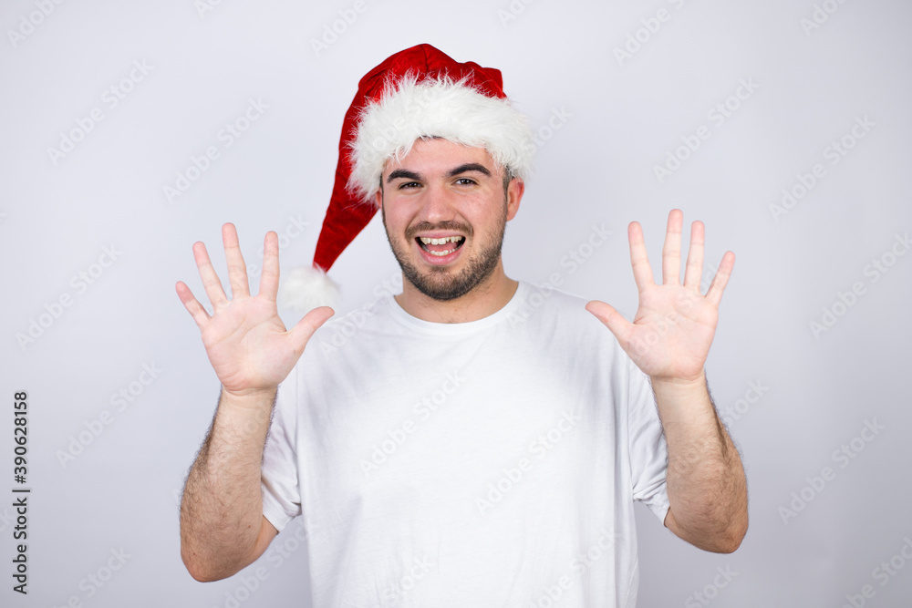 Young handsome man wearing a Santa hat over white background showing and pointing up with fingers number ten while smiling confident and happy Young handsome man wearing a Santa hat over white background showing and pointing up with fingers number ten while smiling confident and happy