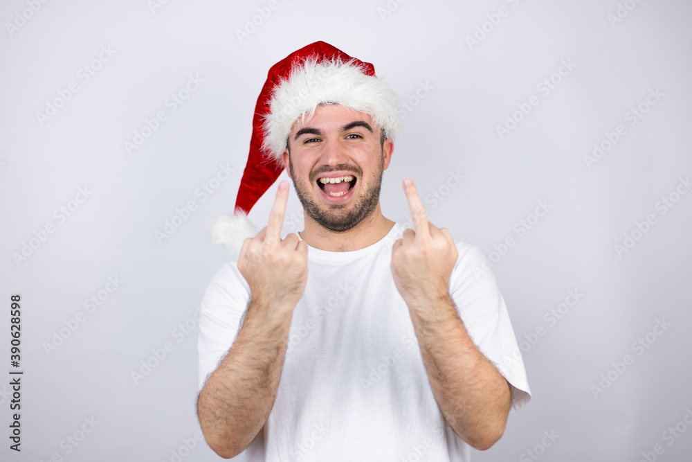 Young handsome man wearing a Santa hat over white background showing middle finger doing fuck you bad expression, provocation and rude attitude. screaming excited Young handsome man wearing a Santa hat over white background showing middle finger doing fuck you bad expression, provocation and rude attitude. screaming excited