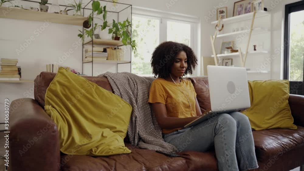 Portrait of smiling african american young woman with earphones typing on laptop while sitting on sofa in living room Portrait of smiling african american young woman with earphones typing on laptop while sitting on sofa in living room