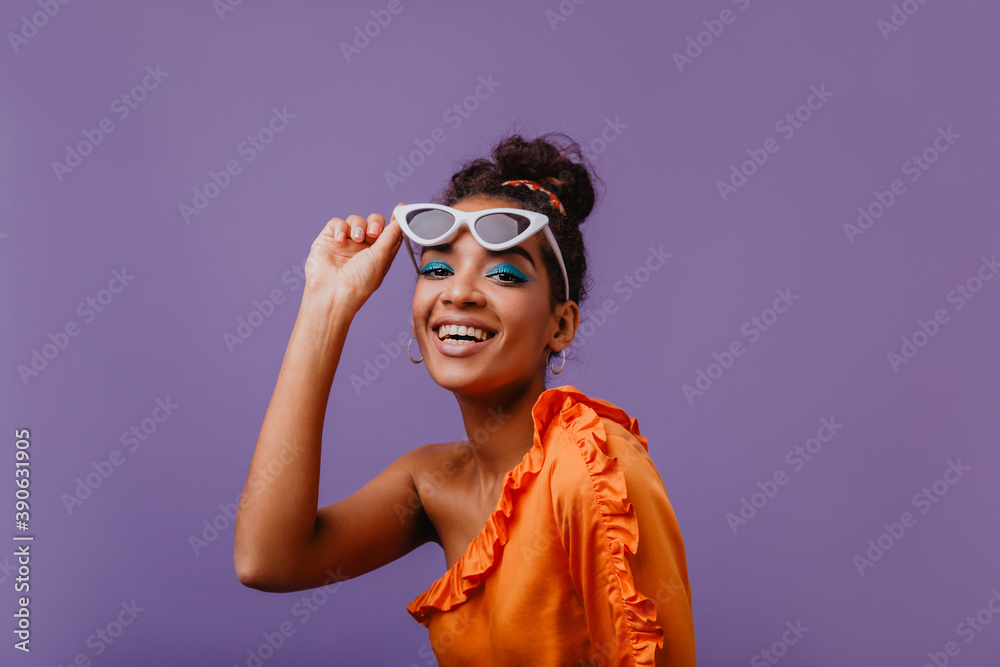 Gorgeous stylish girl with blue makeup posing with sunglasses. Indoor photo of ecstatic african female model sitting on purple background with smile.