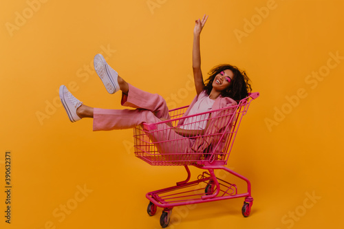 Chilling african girl posing in shopping cart. Indoor photo of emotional young lady in pink clothes. photo
