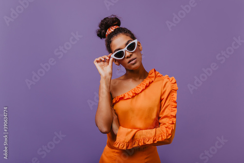 Serious african lady in orange blouse standing in front of purple wall. Indoor photo of enthusiastic curly woman in sunglasses. © Look!