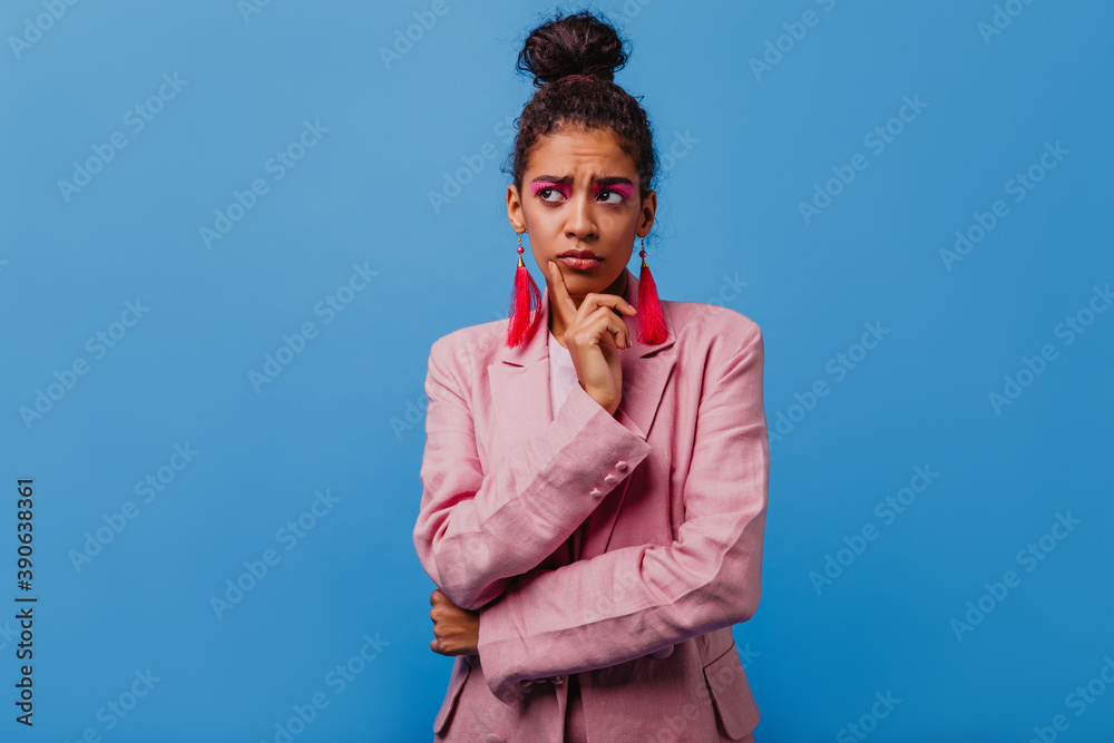 Sad african woman wears pink earrings standing in studio. Upset gorgeous girl in jacket posing on blue background. Sad african woman wears pink earrings standing in studio. Upset gorgeous girl in jacket posing on blue background.